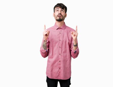 Yakışıklı delikanlı izole arka plan üzerinde pembe gömlek giymiş hayrete ve yukarıya bakıyor ve parmakları ile işaret ve silah kaldırdı şaşırttı.