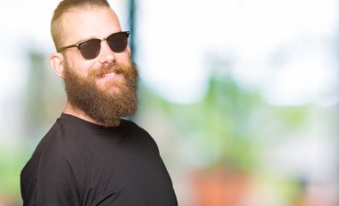 Genç hipster adam uzak tarafına yüzünde, doğal ifade gülümseme ile seyir güneş gözlüğü takıyor. Kendine güvenen gülüyor.