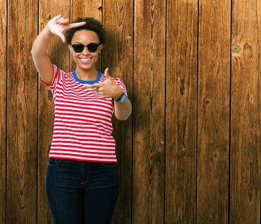 Güzel genç Afro-Amerikan kadın elle yapma çerçeve ve parmakları ile mutlu yüz gülümseyen izole arka plan üzerinde güneş gözlüğü takıyor. Yaratıcılık ve Fotoğrafçılık kavramı.