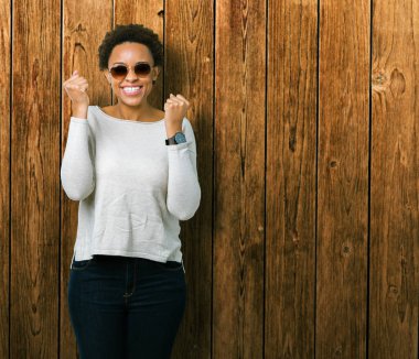 Güzel genç Afro-Amerikan kadın izole arka plan kutlama üzerinde güneş gözlüğü takıyor şaşırttı ve silah ile başarı için yükseltilmiş ve açık gözler hayrete düşürdü. Kazanan kavramı.