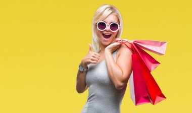 Genç güzel sarışın kadın alışveriş holding alışveriş torbaları satış izole arka yapıyor ok işareti, kocaman bir gülümseme ile mutlu üzerinde parmak ile mükemmel işareti başparmak