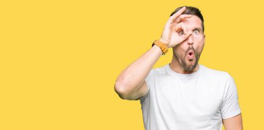 Yakışıklı adam ok jest yapıyor casual beyaz t-shirt giyen sürpriz yüz, göz parmaklarının arasından seyir ile şok. İmansız ifade.