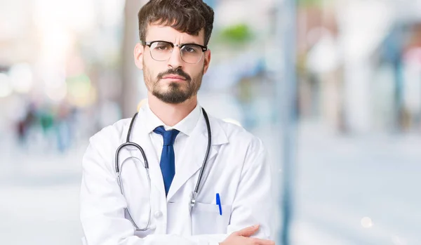 Young doctor man wearing hospital coat over isolated background skeptic and nervous, disapproving expression on face with crossed arms. Negative person.