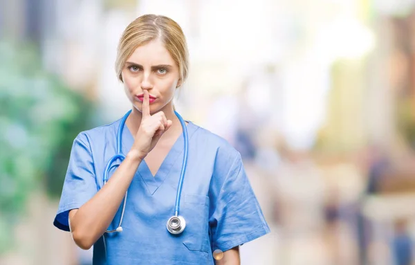 Young beautiful blonde doctor surgeon nurse woman over isolated background asking to be quiet with finger on lips. Silence and secret concept.