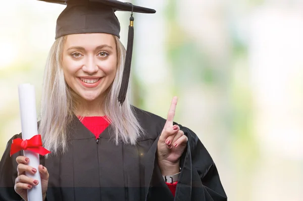 年轻的金发女子穿着研究生制服持有程度在孤立的背景下惊讶的想法或问题指着一个快乐的脸 — 图库照片