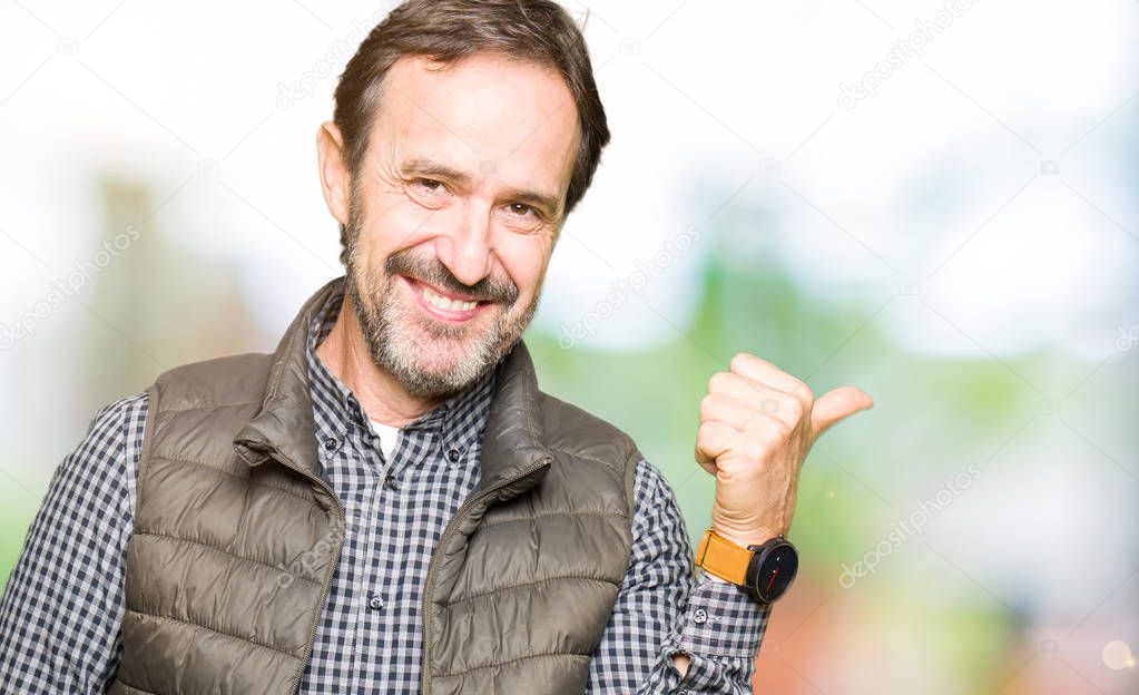 Middle age handsome man wearing winter vest smiling with happy face looking and pointing to the side with thumb up.