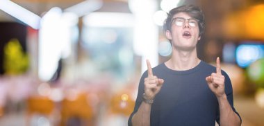 Yakışıklı delikanlı hayrete ve yukarıya bakıyor ve parmakları ile işaret ve silah kaldırdı sürpriz izole arka plan üzerinde gözlük.