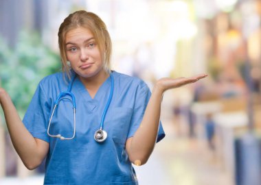 Üzerinde cerrah üniforma giyen genç beyaz doktor kadın arka plan clueless ve şaşkın bir ifadeyle kollar ve eller kaldırdı izole. Şüphe kavramı.