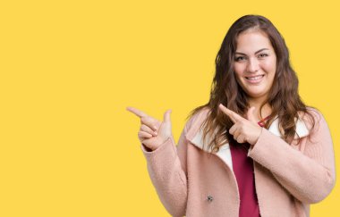 Artı boyutu genç kadın güzel üzerinde kış palto giyiyor gülümseyen ve iki elleri ve parmakları ile tarafına işaret kamera bakarak arka plan izole.