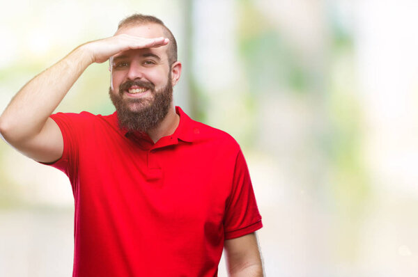 Young caucasian hipster man wearing red shirt over isolated background very happy and smiling looking far away with hand over head. Searching concept.