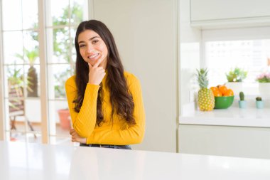 Genç güzel kadın evde beyaz masada emin çapraz kollar ve çeneme kaldırdı el ile gülümseme ile kameraya bakıyor. Pozitif düşünce.