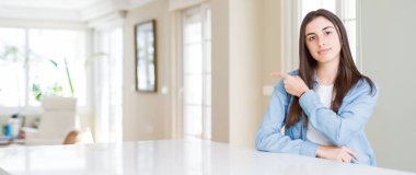 Güzel genç kadın evde beyaz masaya oturan geniş açı resim el parmak ile gösterilen reklam, ciddi ve sakin yüz tarafına işaret.