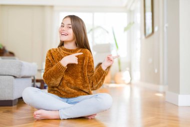 Güzel genç kız çocuk evde katta oturan gülümseyen ve iki elleri ve parmakları ile tarafına işaret kamera bakıyor.