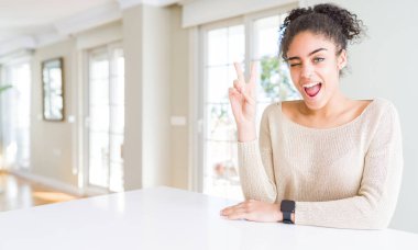 Mutlu yüz zafer işareti yaparken kameraya göz kırpıyor ile evde masaya oturan afro saçlı güzel genç Afro-Amerikan kadın gülümsüyor. İki numaralı.