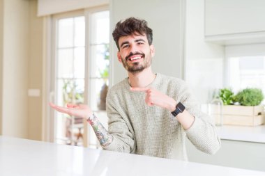 Hayret ve el ile sunulması ve parmak ile işaret ederken kameraya gülümseyen beyaz masada oturan rahat kazak giyen genç adam.