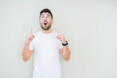 Yakışıklı delikanlı izole arka plan üzerinde rahat beyaz t-shirt giyen hayrete ve yukarıya bakıyor ve parmakları ile işaret ve silah kaldırdı şaşırttı.