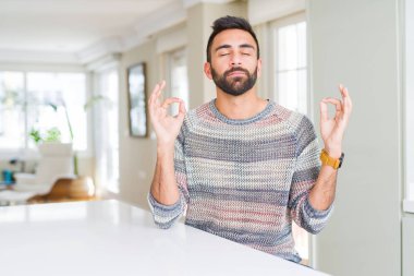 Evde rahat kazak giyen yakışıklı İspanyol adam sakin ol ve gülümseyen gözlerle kapalı parmak jestle meditasyon yapıyor. Yoga kavramı.