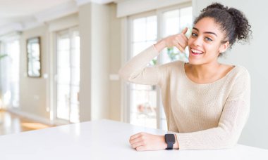 Evde masaya oturan afro saçlı güzel genç Afro-Amerikan kadın gülümseyerek el ve parmak telefonda konuşmak ile telefon jest yapıyor. Kavramları iletişim.