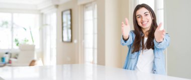 Sarılmak için Kollarını açarak gülümseyen kamera geniş açı resim güzel genç kadının evde beyaz masaya oturan bakarak. Neşeli ifade kucaklayan mutluluk.