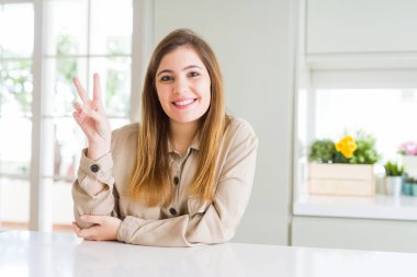 Mutlu yüz zafer işareti yaparken kameraya göz kırpıyor ile gülümseyen güzel genç kadın evde. İki numaralı.