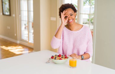 Sabahları sağlıklı kahvaltı evde olan genç Afro-Amerikan kadın gülümsüyor, parmakları ile mutlu yüz üzerinden seyir göz el ile ok jest yapıyor.