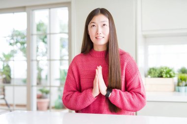 Güzel Asyalı kadın yalvarıyor ve umut ifade yüzünde çok duygusal ve endişe ile birlikte elleriyle dua beyaz masa üzerinde pembe kazak giyiyor. Bağışlanmak. Din kavramı.