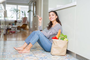 Kameraya bakarak yan el ve parmak ile işaret yüzünde kocaman bir gülümseme ile mutfakta taze yiyecek dolu kese kağıdı ile oturan genç kadın.