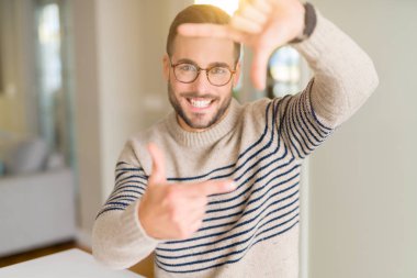 Evde gözlüklü genç yakışıklı adam elleriyle yapım çerçeve ve parmakları ile mutlu yüz gülümsemeye. Yaratıcılık ve Fotoğrafçılık kavramı.