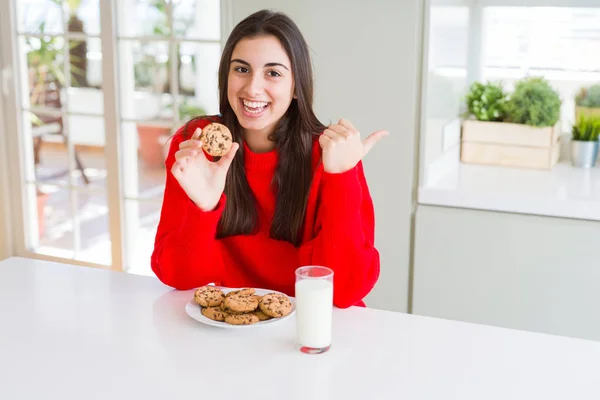 Beautiful young woman eating chocolate chips cookies and glass of milk pointing and showing with thumb up to the side with happy face smiling
