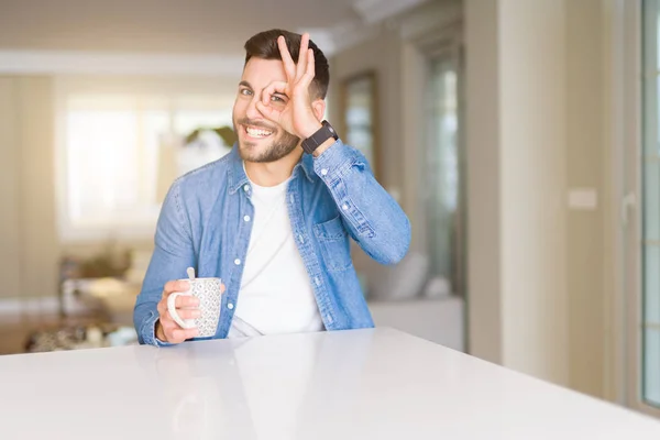 Young handsome man drinking a cup of coffee at home with happy face smiling doing ok sign with hand on eye looking through fingers