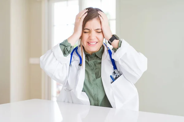 Beautiful young doctor woman wearing medical coat and stethoscope suffering from headache desperate and stressed because pain and migraine. Hands on head.