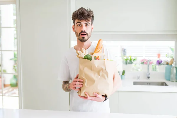 Young man holding paper bag of fresh groceries from the supermarket scared in shock with a surprise face, afraid and excited with fear expression