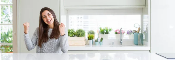 stock image Wide angle picture of beautiful young woman sitting on white table at home celebrating surprised and amazed for success with arms raised and open eyes. Winner concept.