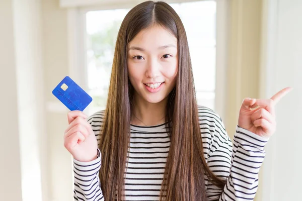 Beautiful Asian woman holding credit card very happy pointing with hand and finger to the side