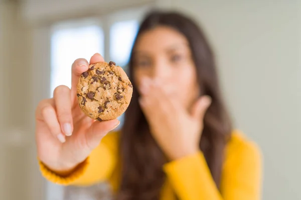 Young woman eating chocolate chips cookies at home cover mouth with hand shocked with shame for mistake, expression of fear, scared in silence, secret concept