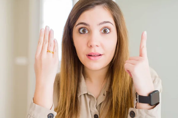 Beautiful young woman showing alliance ring on hand surprised with an idea or question pointing finger with happy face, number one