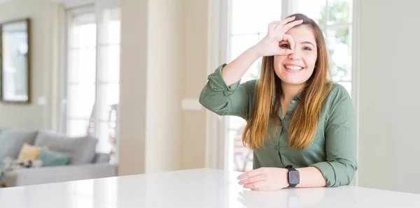 Beautiful young woman at home doing ok gesture with hand smiling, eye looking through fingers with happy face.