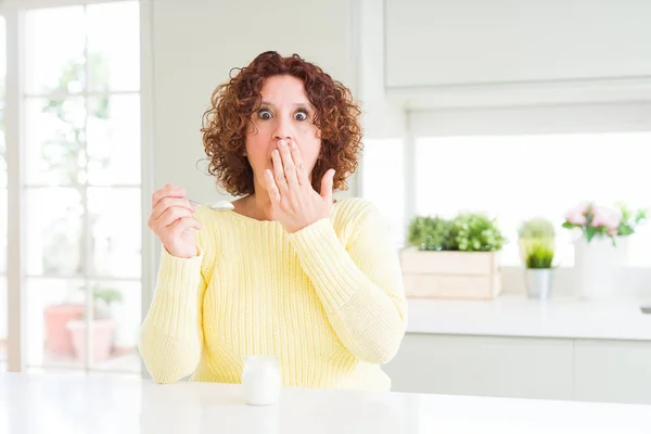 Senior woman eating a healthy natural yogurt at home cover mouth with hand shocked with shame for mistake, expression of fear, scared in silence, secret concept