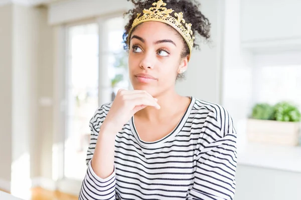 Young african american girl wearing golden queen crown on head with hand on chin thinking about question, pensive expression. Smiling with thoughtful face. Doubt concept.