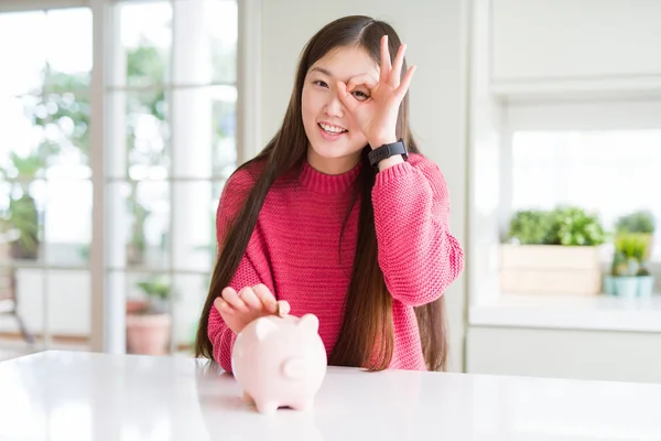 美丽的亚洲女人把一枚硬币在小猪袋里 快乐的脸微笑做确定的标志 用手在眼睛上看着通过手指 — 图库照片