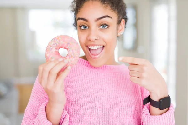 年轻的非洲裔美国女孩吃甜粉红色甜甜圈非常高兴用手和手指指着 — 图库照片