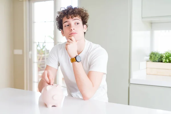 Young man investing money using piggy bank at home serious face thinking about question, very confused idea