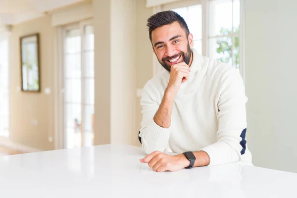 Handsome hispanic man wearing casual white sweater at home looking confident at the camera with smile with crossed arms and hand raised on chin. Thinking positive.