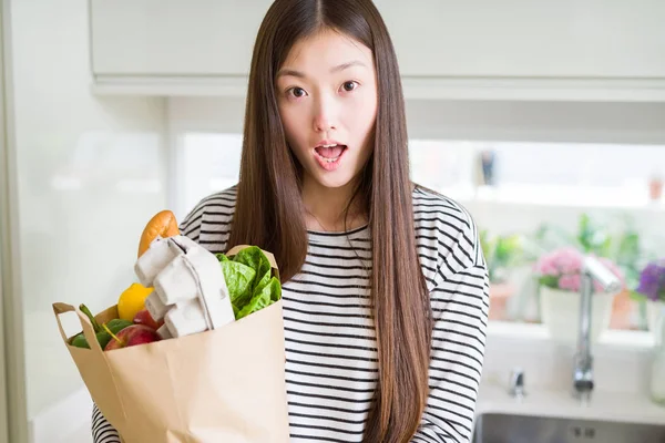 Beautiful Asian woman holding paper bag of fresh groceries scared in shock with a surprise face, afraid and excited with fear expression