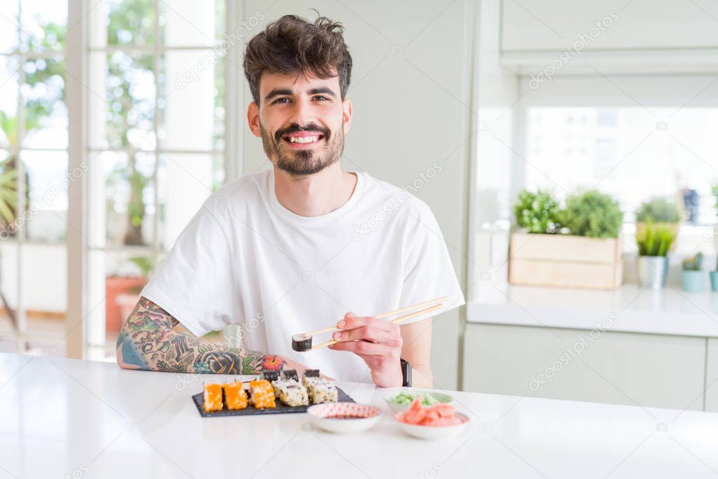 Young man eating asian sushi from home delivery with a happy face standing and smiling with a confident smile showing teeth