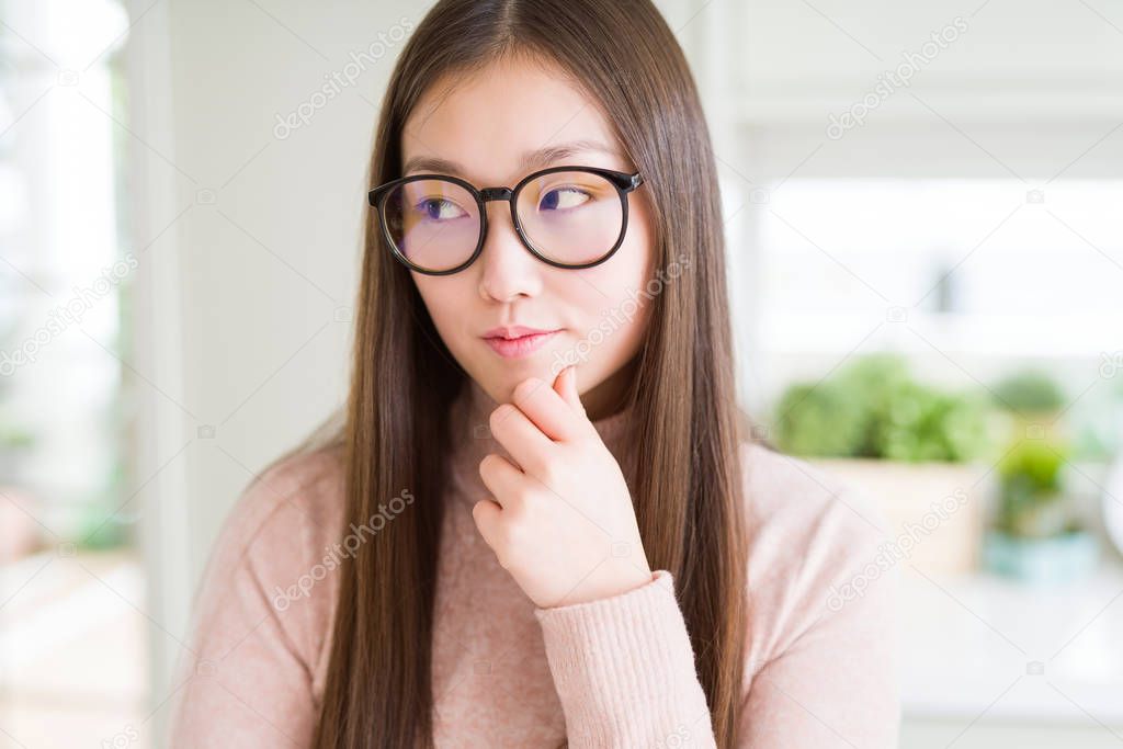 Beautiful Asian woman wearing glasses with hand on chin thinking about question, pensive expression. Smiling with thoughtful face. Doubt concept.
