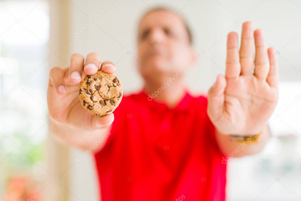 Middle age man eating chocolate chips cookies at home with open hand doing stop sign with serious and confident expression, defense gesture