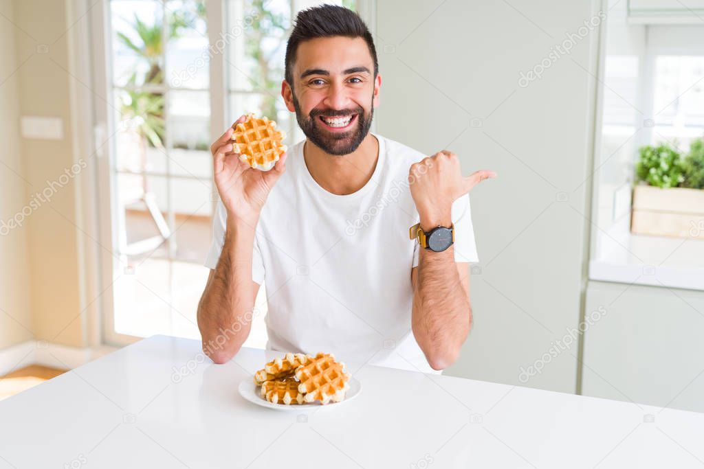 Handsome hispanic man eating sweet belgian waffle pastry pointing and showing with thumb up to the side with happy face smiling