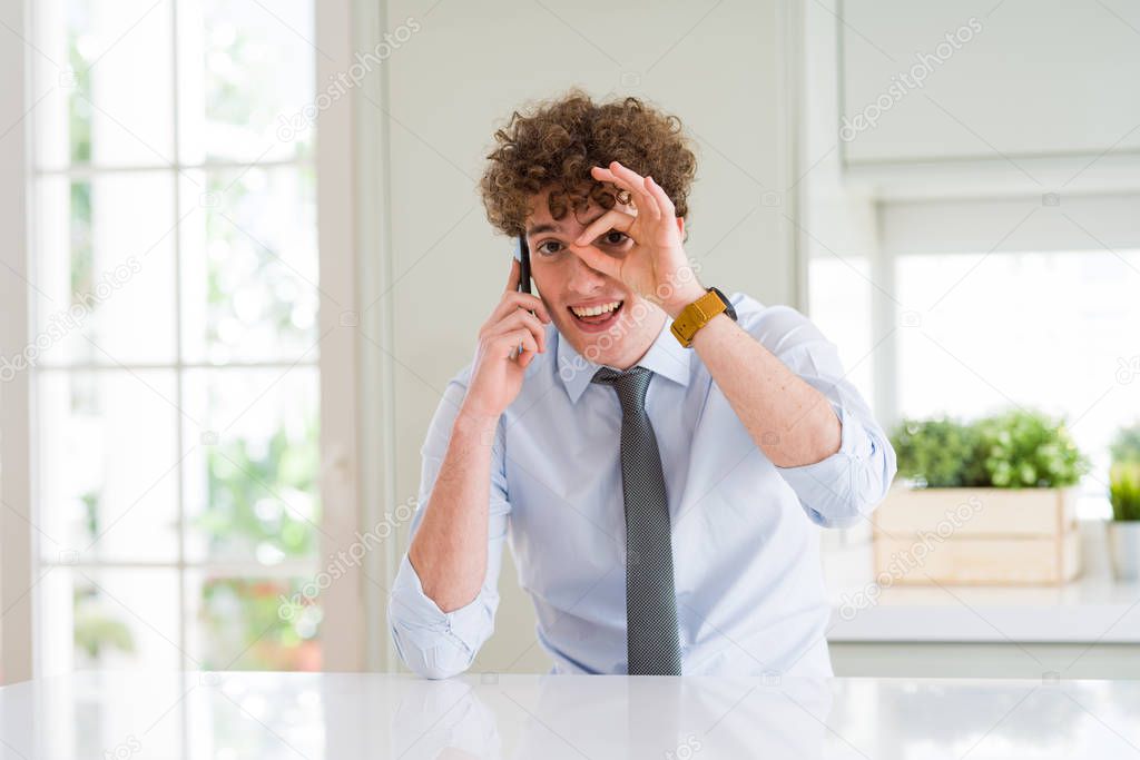 Young business man talking on smartphone at the office with happy face smiling doing ok sign with hand on eye looking through fingers