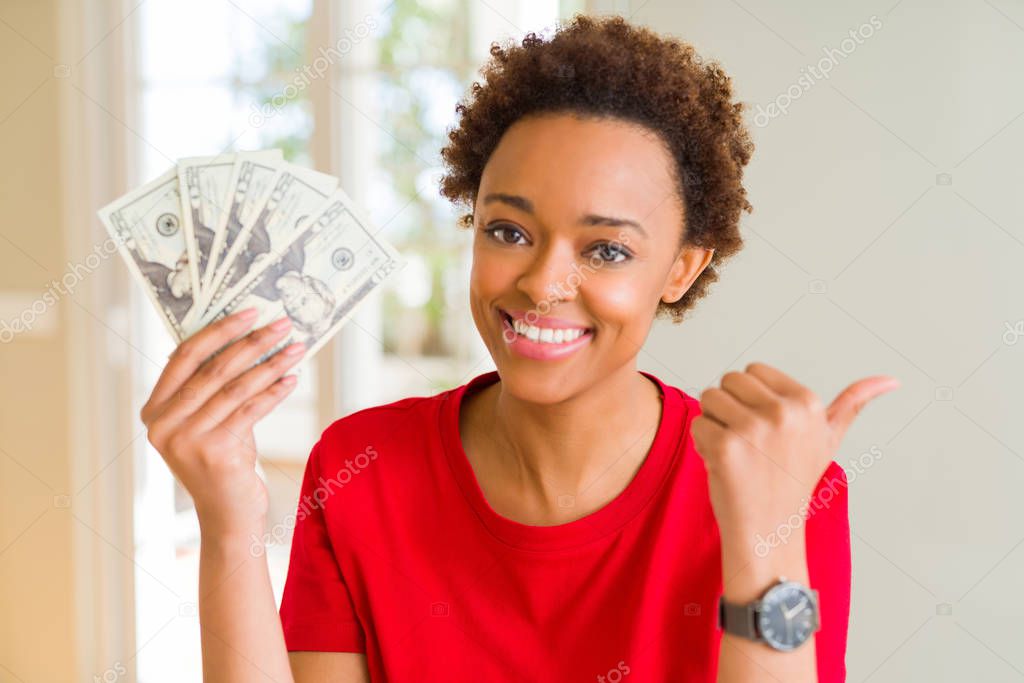 Young african american woman holding bank notes of twenty dollars pointing and showing with thumb up to the side with happy face smiling